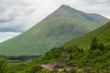 Gray clouds touch the grassy top of mountain under which the railway line winds