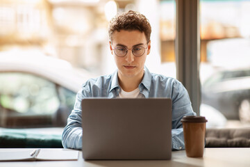 A focused young man with curly hair and glasses intently uses a laptop in a well-lit cafe