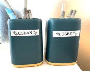 Pen containers sitting on an office counter top. One container is for clean unused pens, and the other is a container for dirty or used pens. 