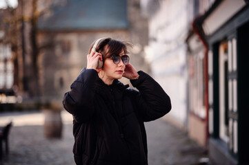 Fototapeta premium A chic adult woman adjusts her over-ear headphones, her expression serene. She's clad in a black jacket, accented with hoop earrings and sunglasses, in an old-town setting