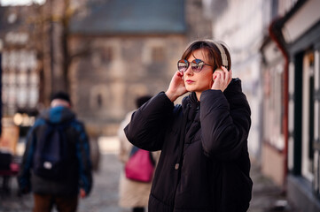 A chic adult woman adjusts her over-ear headphones, her expression serene. She's clad in a black...