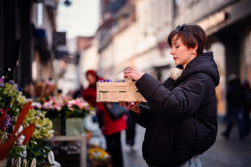 A stylish woman with headphones examines a crate of colorful flowers at a vibrant city market,...