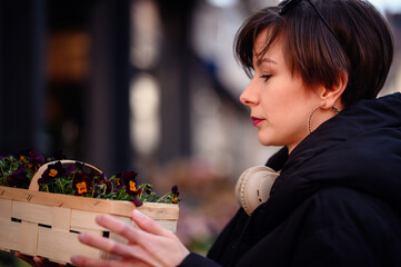A concentrated woman carefully chooses among deep purple pansies at a lively outdoor market, her headphones hinting at a melodic shopping experience
