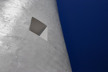 On the curved exterior of a lighthouse a square window looks out on a clear blue sky.