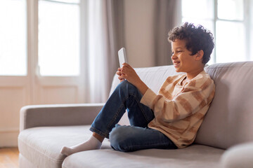 Cheerful black boy sitting relaxed on sofa, deeply engaged in his smartphone