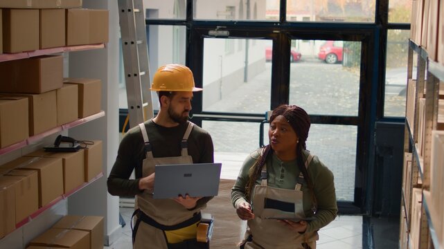 Meticulous employees preparing warehouse orders for delivery, using laptop and tablet to verify shipping informations, crosschecking online data with merchandise labels in storage facility