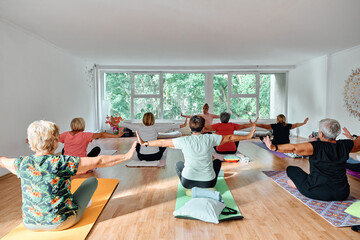 A group of senior women engage in various yoga exercises, including neck, back, and leg stretches, under the guidance of a trainer in a sunlit space, promoting well-being and harmony