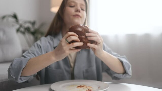 Portrait of funny happy young woman eating burger with great pleasure while ketchup flows down the hands at home Excited hungry female biting hamburger enjoying fast junk food indoors