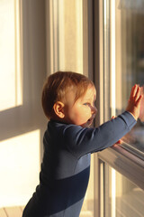 Fototapeta na wymiar A cute little boy stands by the window and looks at the street. Hard light, sunrise or sunset