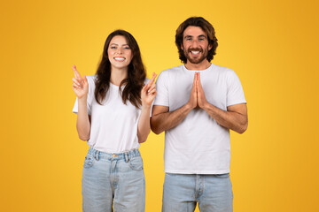 Hopeful young woman with crossed fingers and cheerful man with palms together in a gesture of...