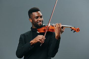 Elegant African American man in black suit playing violin against gray background for music and...