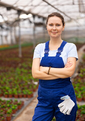 young positive woman in overalls poses with arms crossed on chest next to plantation of young plants in greenhouse. plant-growing farm worker © JackF