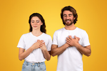 Grateful young couple with eyes closed and hands on heart, feeling thankful and serene in plain white t-shirts