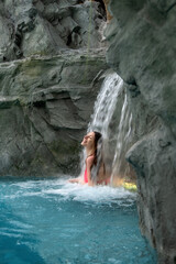 young sexy blond woman in fuchsia pink bikini enjoys the falling water of the waterfall in the spa wellness pool