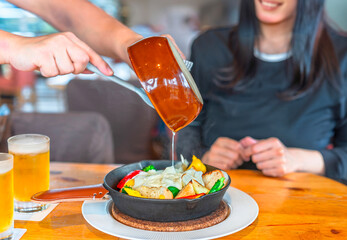 Focus on the hands of a restaurant waiter holding a ceramic frying pan and pouring melted cheese...