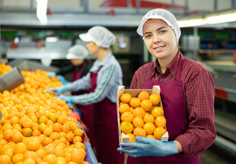 Portrait of cheerful young girl worker standing with box of selected tangerines at fruit sorting...
