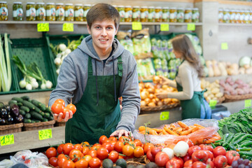 Smiling young salesman putting tomatoes in vegetable basket in large grocery store