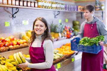 Happy young shop assistant setting bunch of bananas on a counter in big greengrocery