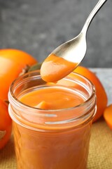 Taking delicious persimmon jam with spoon from glass jar on table, closeup