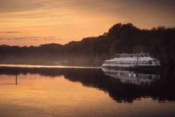 Ship is sailing on calm river near forest at golden sunset with reflections on the water