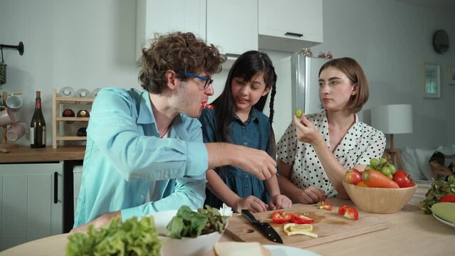 Attractive caucasian father and mother spend time together to cook fresh meal with asian daughter. Dad and mom preparing fresh vegetable for making salad at kitchen and eating vegetable. Pedagogy.