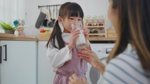 Asian little daughter drinking a glass of milk with mother in kitchen. 