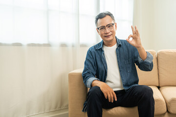 Handsome asian mature old man sitting on sofa couch in living room at home. Happy Portrait of cheerful smiling senior asian man relax in the house. Mature People and lifestyle