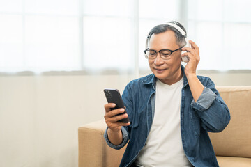 Mature lifestyle relaxing on sofa in the morning. Happy Asian senior old man listening music with smartphone put on headphone by streaming music online. Choosing song by cell phone