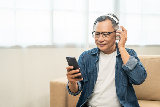 Mature lifestyle relaxing on sofa in the morning. Happy Asian senior old man listening music with smartphone put on headphone by streaming music online. Choosing song by cell phone
