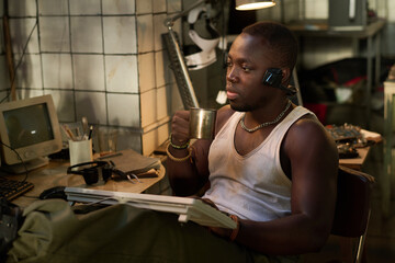 Medium shot of young African American programmer sitting at workplace in post-apocalyptic shelter working on old computer and drinking tea