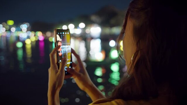 Asian woman takes photos of night river reflections in Hoi An, Vietnam with blurred background