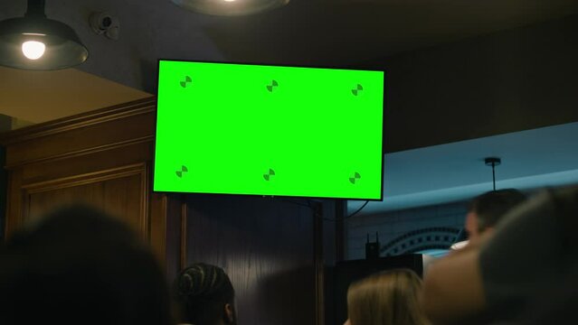 Close up shot of TV on wall with displayed live match broadcast with Chromakey Green Screen. Crowd of fans gathered together to cheer team playing in tournament. Group of friends sit in sports pub.