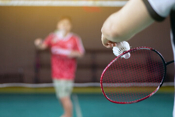 Badminton player holds racket and white cream shuttlecock in front of the net before serving it to another player in opposite side of the court, soft focus.