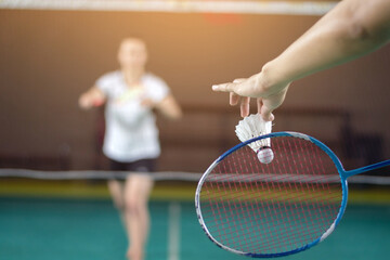 Badminton player holds racket and white cream shuttlecock in front of the net before serving it to another player in opposite side of the court, soft focus.