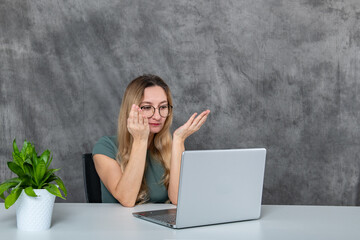 Adorable girl in glasses and gray dress pulling faces by the laptop next to a green plant