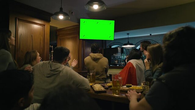 Group of multi ethnic friends sit in sports pub and watch live broadcast on TV Green Screen, chromakey. People gathered to cheer team playing in tournament. Crowd of fans support club during football