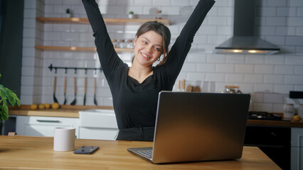 Joyful beautiful woman sitting in the kitchen checking emails on laptop read notice receive good...