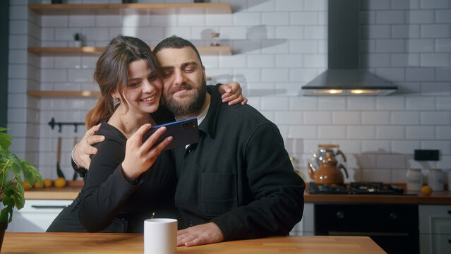 Cozy couple sitting in the kitchen taking selfie photos with smartphone smiling, having fun, having great time	
