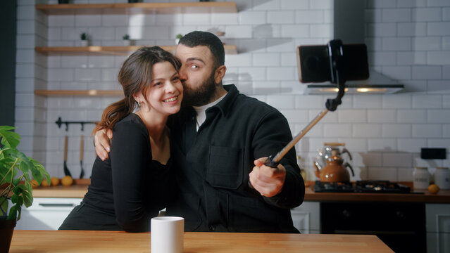 Happy couple sitting in the kitchen taking photos with smartphone on selfie stick laughing, having fun	

