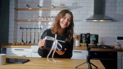 Female young influencer vlogger woman sit in the kitchen holding gift box recording video blog on...