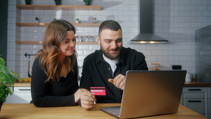 Young couple sit in the kitchen picking goods to buy man enter credit card number on laptop for...