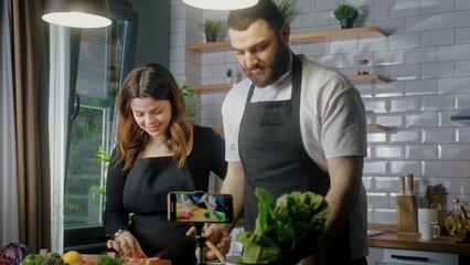 Young couple in an apron standing in kitchen records on smartphone food videoblog. Couple cutting vegetables, preparing healthy a salad. Blogging, healthy lifestyle, cooking master class	
