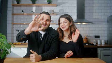 Happy young couple sit in the kitchen make video call looking at camera wave hands greeting,...