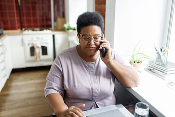 Woman talking on phone sitting at kitchen table. Plus size african american aged female having conversation on cellphone with her husband. Black overweight grandma in glasses calling her grandchild - 756973728