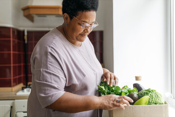 Side view of plus size black grandmother in glasses picking up vegetables from carton box of food...