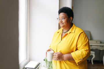 Side view of charming elderly plus size african american woman in yellow shirt holding white cup of...