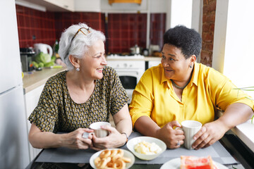 Two aged ladies of diverse ethnicity sitting at kitchen table drinking coffee, sharing gossips and rumors, discussing news, looking at each other with laughing faces, enjoying spending time together - 756974171