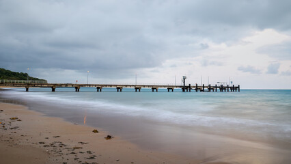 Dawn on an overcast day with a view of a fishing and small boat jetty on a tropical beach with coastal rainforest and smooth sea because of long exposure photography at Mission Beach in Australia.