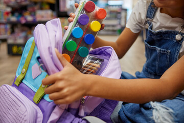 Portrait of lovely smart girl putting paints kit into schoolbag