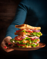 Woman palm of hand holding a plate with huge homemade juicy sandwich made of layers with sliced loaf of bread, green lettuce, fresh tomato, onion rings, cucumber and ham used as fast food for lunch